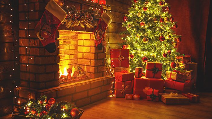 Green Christmas tree with lights beside an open red brick wall fireplace. Stacked presents in red wrapping around the tree and two red stocking hanging above the fireplace.