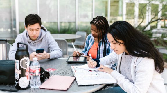 three post-secondary students sitting at a round table outside and studying.