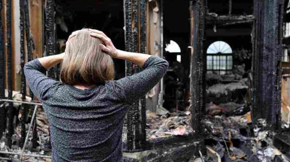 A distressed woman stands in front of a burned-down home, holding her head in shock after a disaster.
