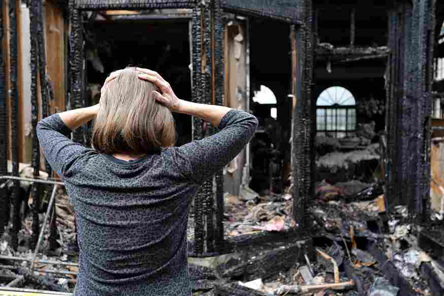 A distressed woman stands in front of a burned-down home, holding her head in shock after a disaster.
