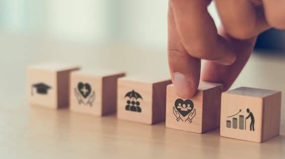 A hand places a wooden block with a heart and group of people icon into a row of blocks that also feature icons for education (graduation cap), health (heart with cross), insurance (umbrella over people), and retirement savings (figure on rising coins), symbolizing personal well-being and life essentials.