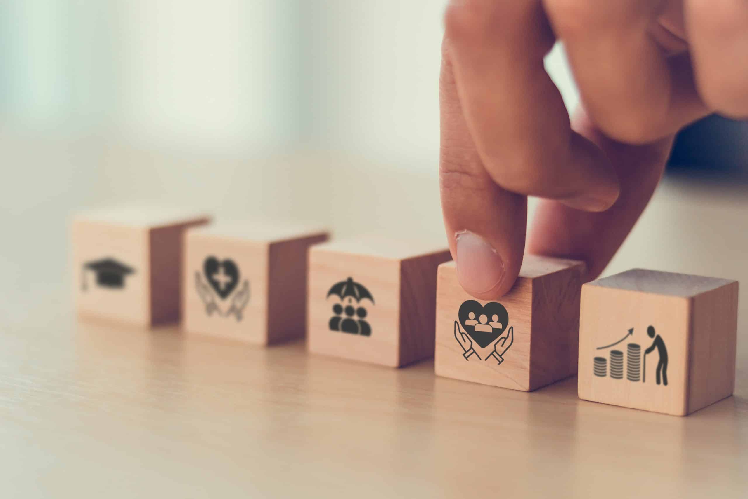 A hand places a wooden block with a heart and group of people icon into a row of blocks that also feature icons for education (graduation cap), health (heart with cross), insurance (umbrella over people), and retirement savings (figure on rising coins), symbolizing personal well-being and life essentials.