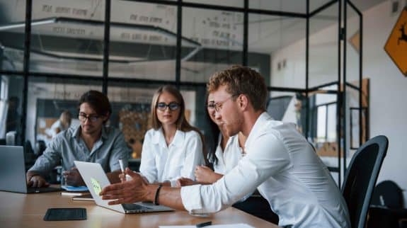 Business team collaborating in a modern glass-walled office, discussing ideas around a laptop during a meeting.