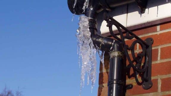 Frozen gutter and downspout with hanging icicles on a brick house during winter, showing the risk of ice buildup and frozen drainage.