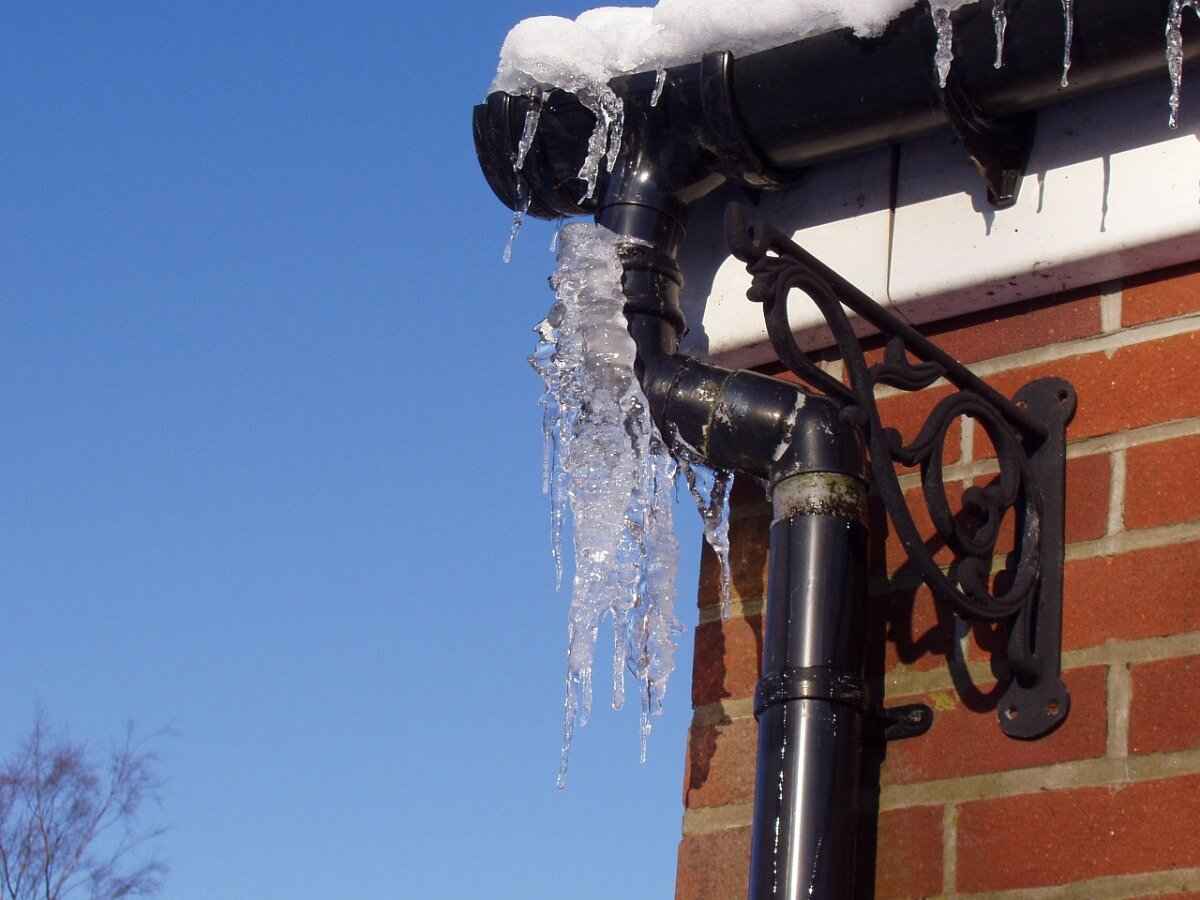 Frozen gutter and downspout with hanging icicles on a brick house during winter, showing the risk of ice buildup and frozen drainage.