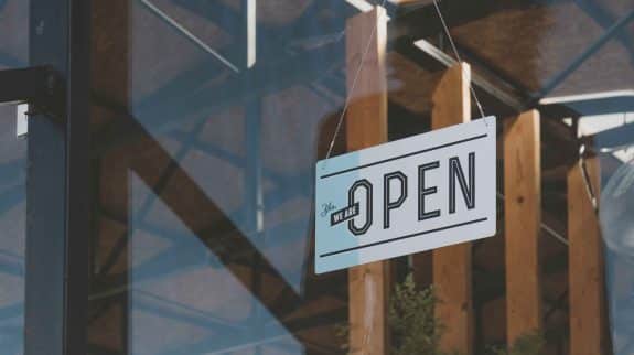 A modern white rectangular sign with black text saying "Yes We Are Open" hanging on a glass door or window of a retail store with a blurred wooden interior background.