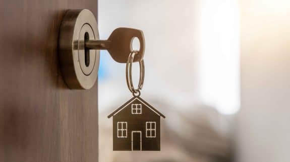 Close-up of a house key with a metal house-shaped keychain in a door lock, representing real estate ownership and home security.