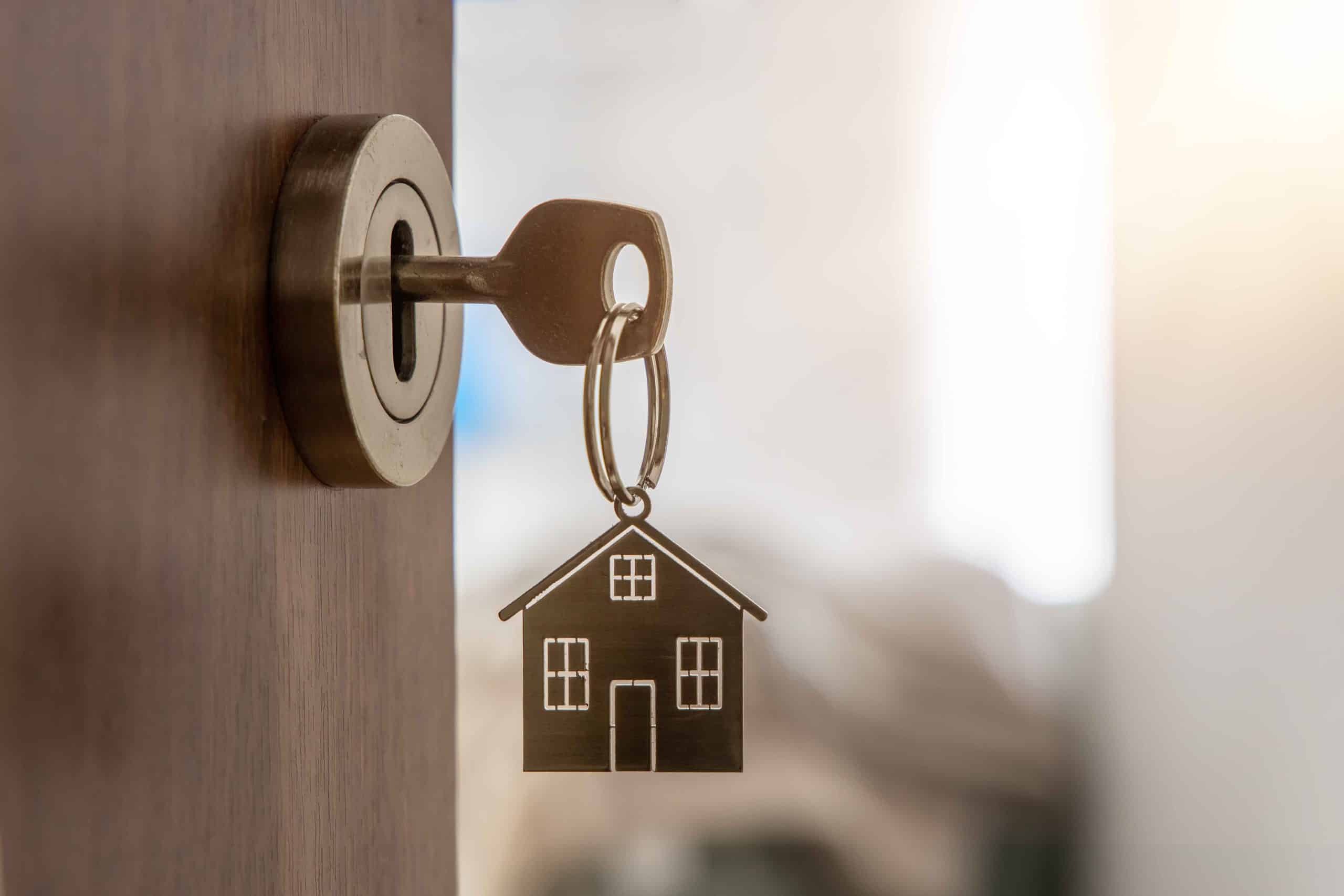 Close-up of a house key with a metal house-shaped keychain in a door lock, representing real estate ownership and home security.