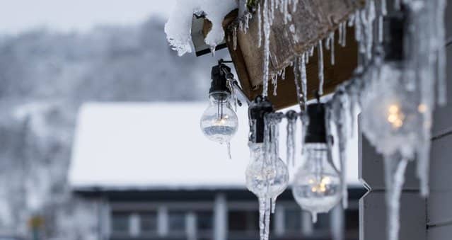 Warm outdoor string lights covered in frozen icicles and snow hanging from a wooden roof with a blurred winter cottage background.