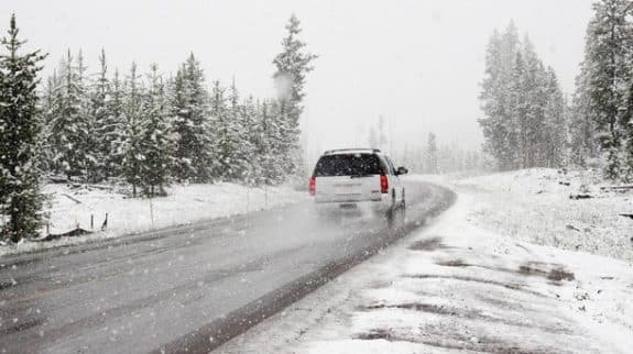 A white SUV driving on a wet, winding road through a snow-covered pine forest during a winter snowstorm.