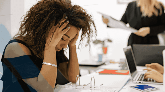 Stressed businesswoman holding her head while reviewing documents at office desk with coworkers in background, representing workplace burnout and deadline pressure.