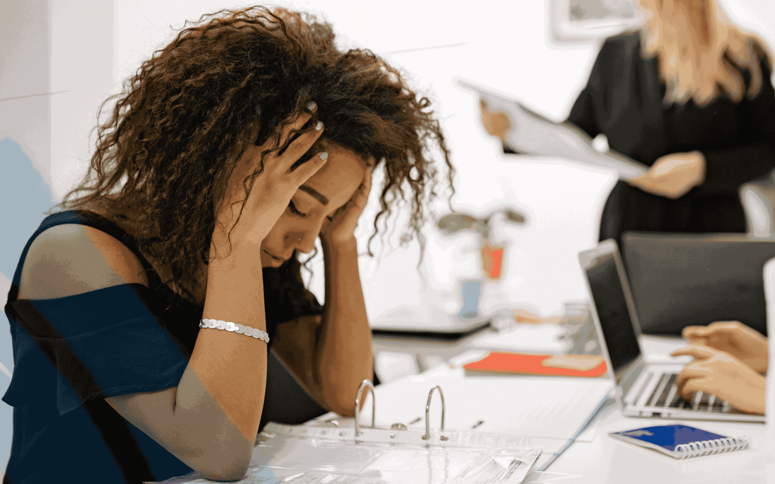 Stressed businesswoman holding her head while reviewing documents at office desk with coworkers in background, representing workplace burnout and deadline pressure.