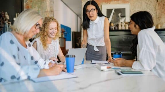 Four women professionals collaborating at a conference table in a modern office, reviewing notes and working together on a laptop during a team meeting.