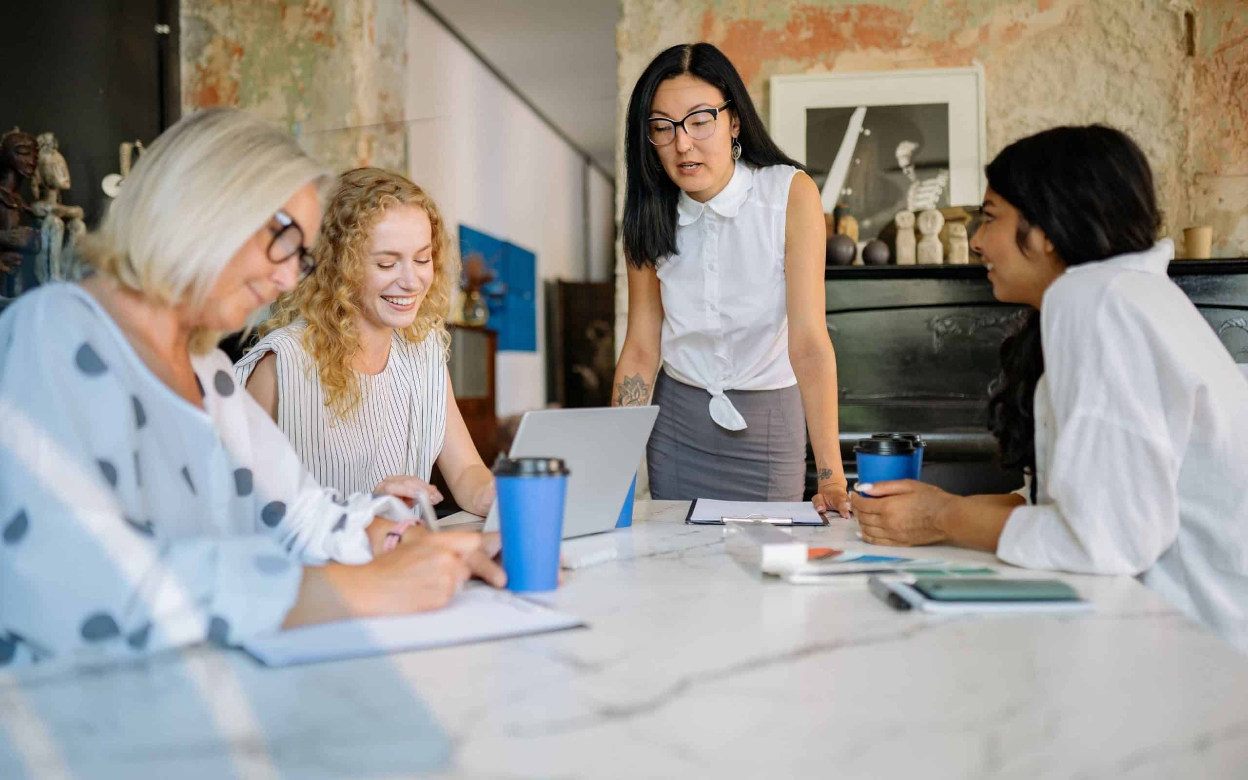 Four women professionals collaborating at a conference table in a modern office, reviewing notes and working together on a laptop during a team meeting.