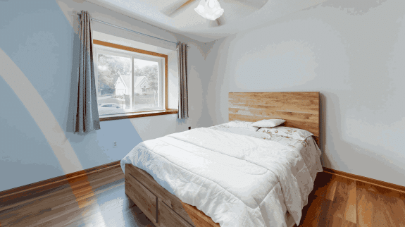 Minimalist bedroom with wooden bed frame, white bedding, hardwood floors, gray curtains, and a sunlit window creating a bright modern interior.