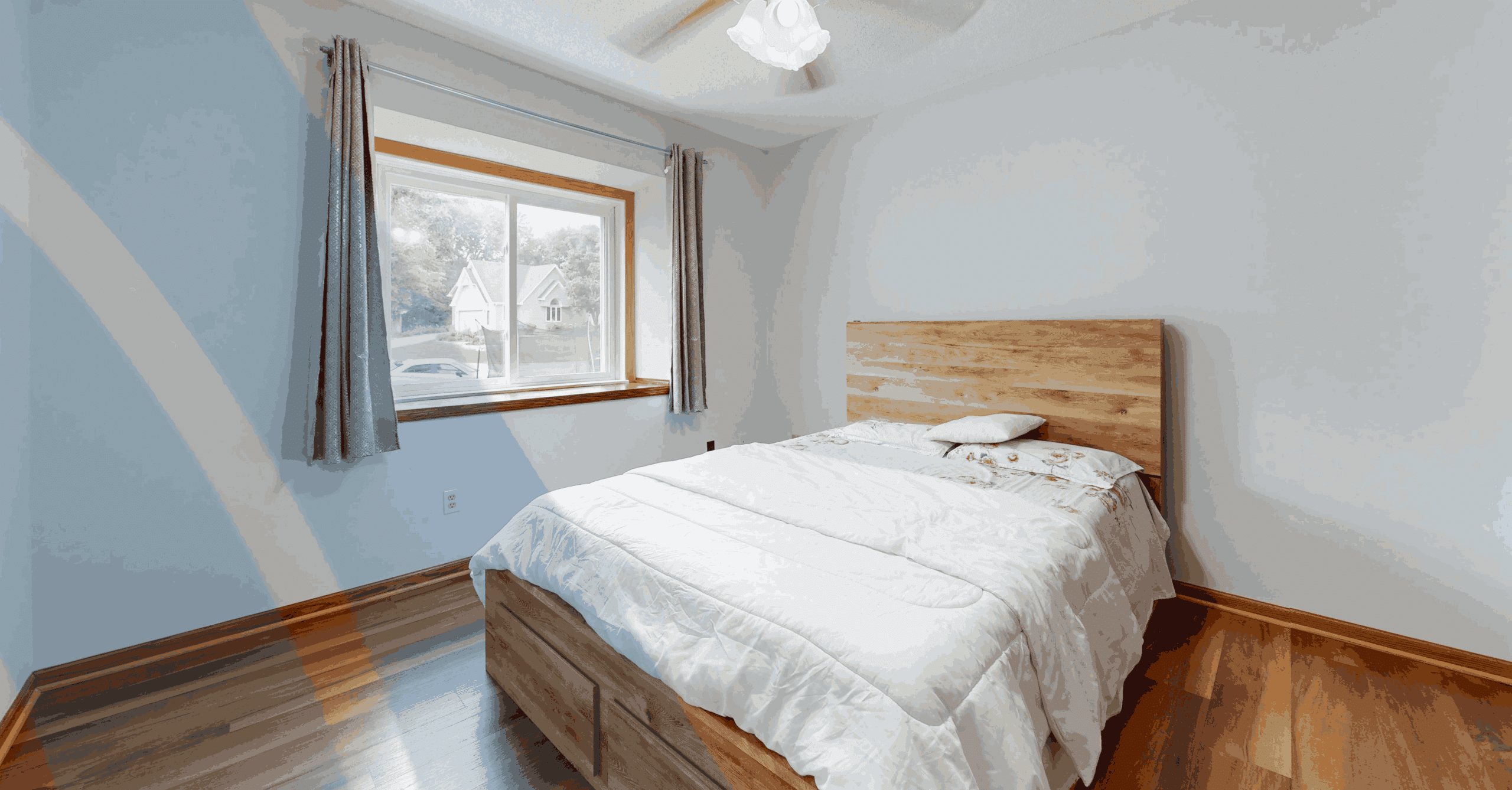 Minimalist bedroom with wooden bed frame, white bedding, hardwood floors, gray curtains, and a sunlit window creating a bright modern interior.