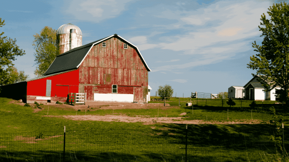 Weathered red barn with metal silo on a rural countryside farm surrounded by green grass, trees, and small white farm buildings under a blue sky