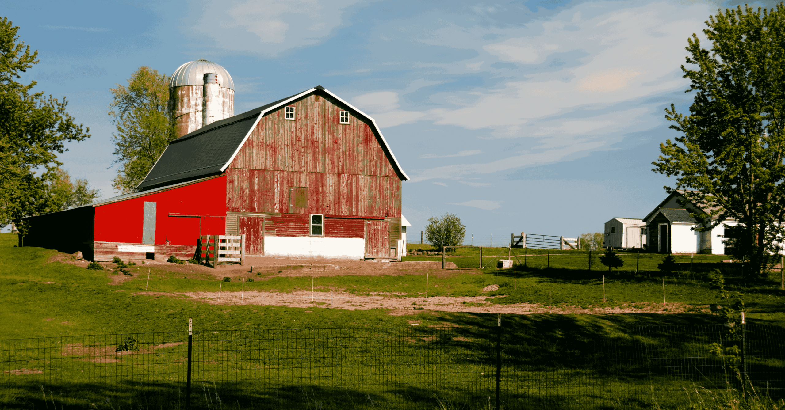 Weathered red barn with metal silo on a rural countryside farm surrounded by green grass, trees, and small white farm buildings under a blue sky