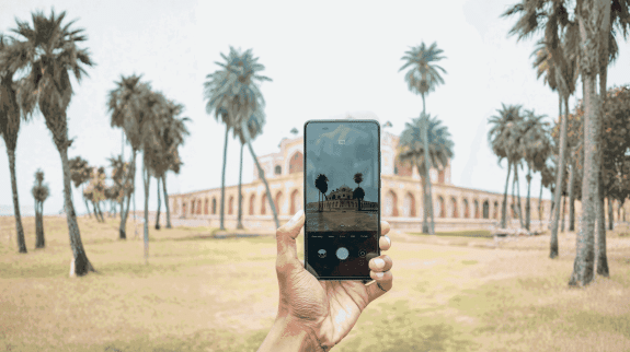 Hand holding smartphone taking a photo of a historic monument with palm trees in a park-like setting.