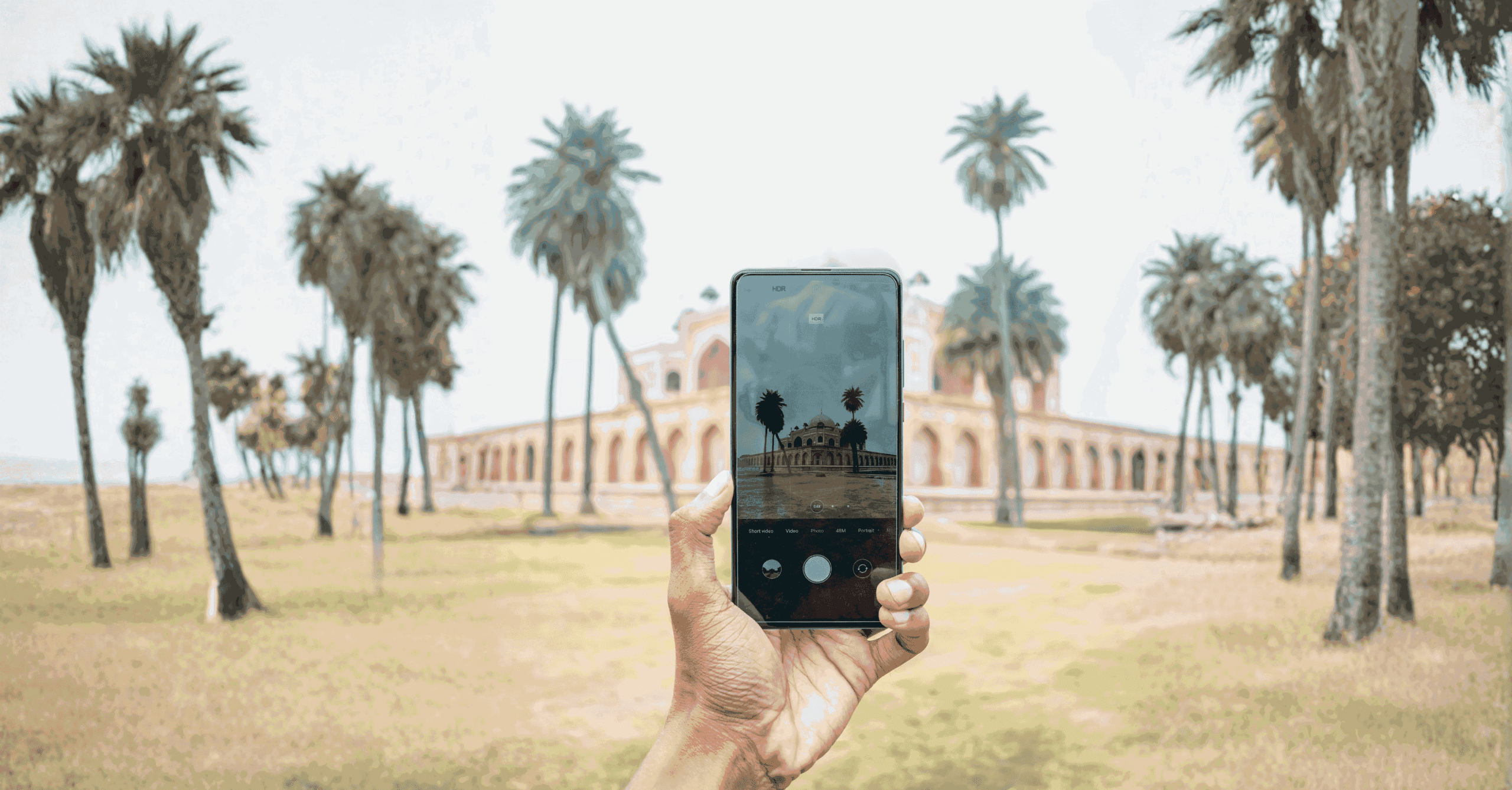 Hand holding smartphone taking a photo of a historic monument with palm trees in a park-like setting.