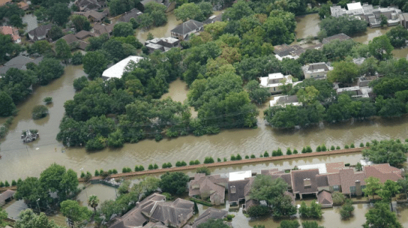 Aerial view of a residential neighborhood affected by river flooding, with brown floodwater surrounding houses, roads, and trees after heavy rainfall.