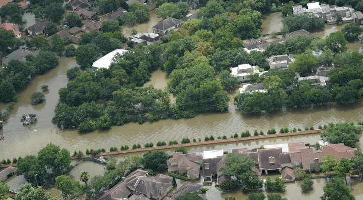 Aerial view of a residential neighborhood affected by river flooding, with brown floodwater surrounding houses, roads, and trees after heavy rainfall.