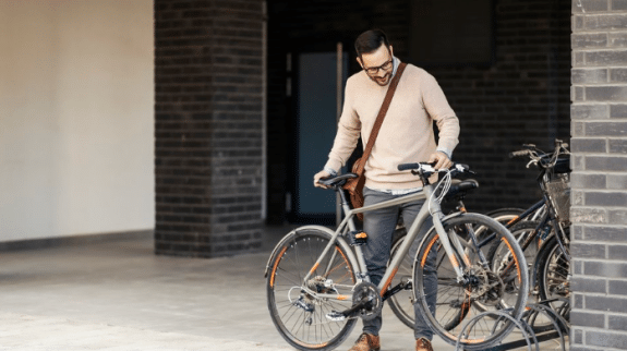 Person standing with a silver bicycle near a bike rack outside a modern brick building in an urban setting