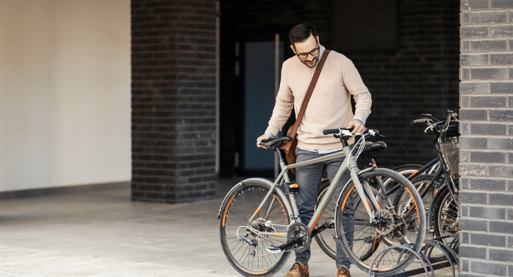 Person standing with a silver bicycle near a bike rack outside a modern brick building in an urban setting