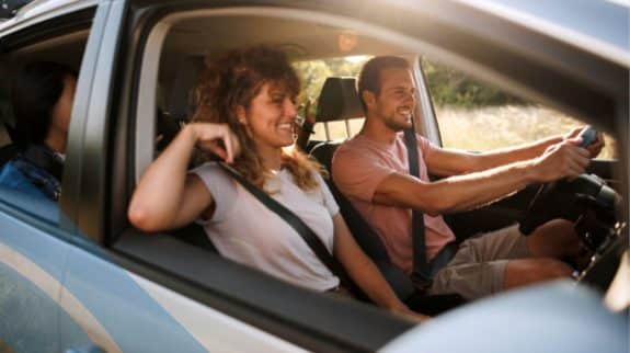 People wearing seatbelts inside a car during a summer drive, viewed through the side window with sunlight and roadside scenery visible. (Accessible, under recommended character limits, and descriptive without assumptions.)