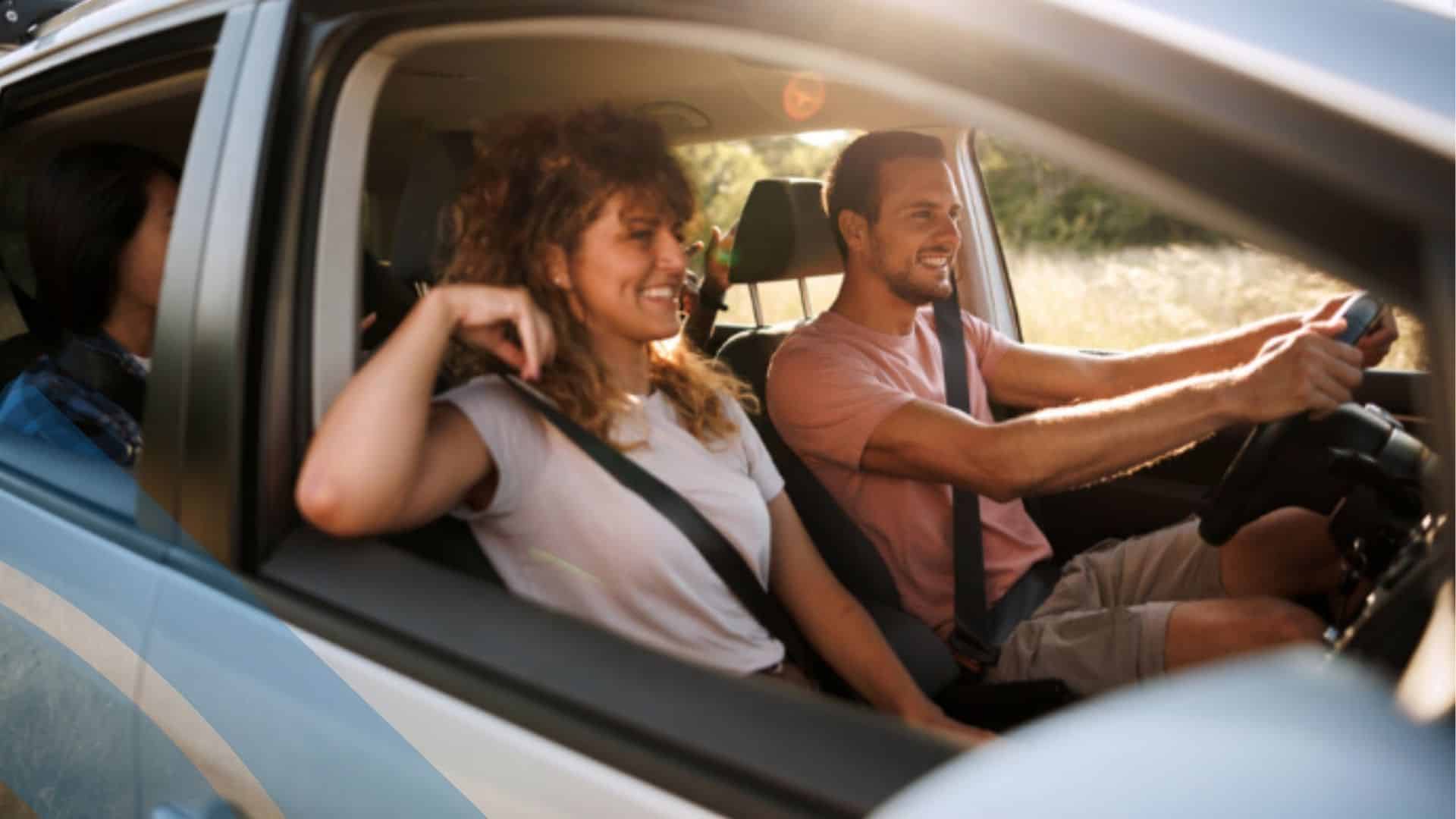 People wearing seatbelts inside a car during a summer drive, viewed through the side window with sunlight and roadside scenery visible. (Accessible, under recommended character limits, and descriptive without assumptions.)