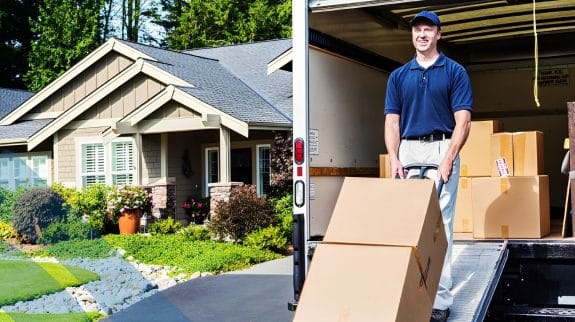 Split image of a suburban residential house exterior and a professional mover lifting a cardboard box from a moving truck during a home relocation.