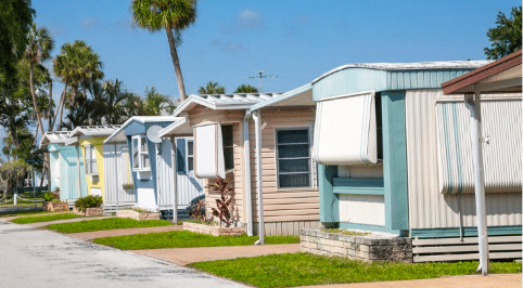 Row of colorful manufactured homes with awnings and manicured lawns in a sunny mobile home park surrounded by palm trees.
