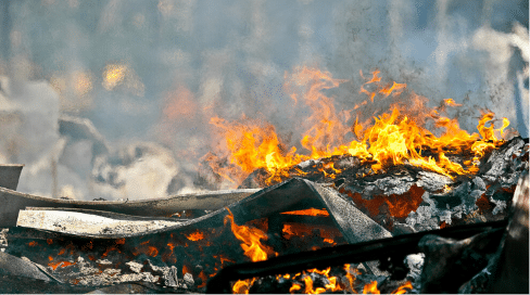 Large fire burning wood and debris with bright orange flames, charred materials, and thick smoke in the background.