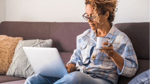 Person sitting on a sofa using a laptop and holding a coffee mug in a cozy home environment.