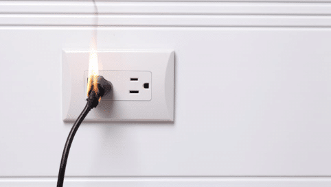Power cord plugged into a wall outlet catching fire with visible flames and smoke, illustrating an electrical fire hazard in a residential home.